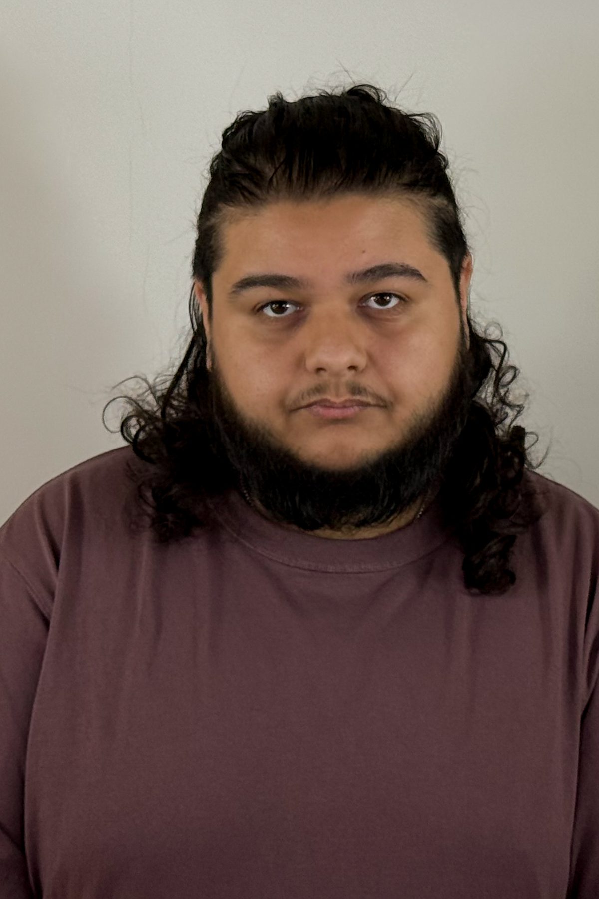 Portrait studio d’Amine Tazit, cheveux longs bouclés, barbe, t-shirt bordeaux, regard neutre face à l’objectif.